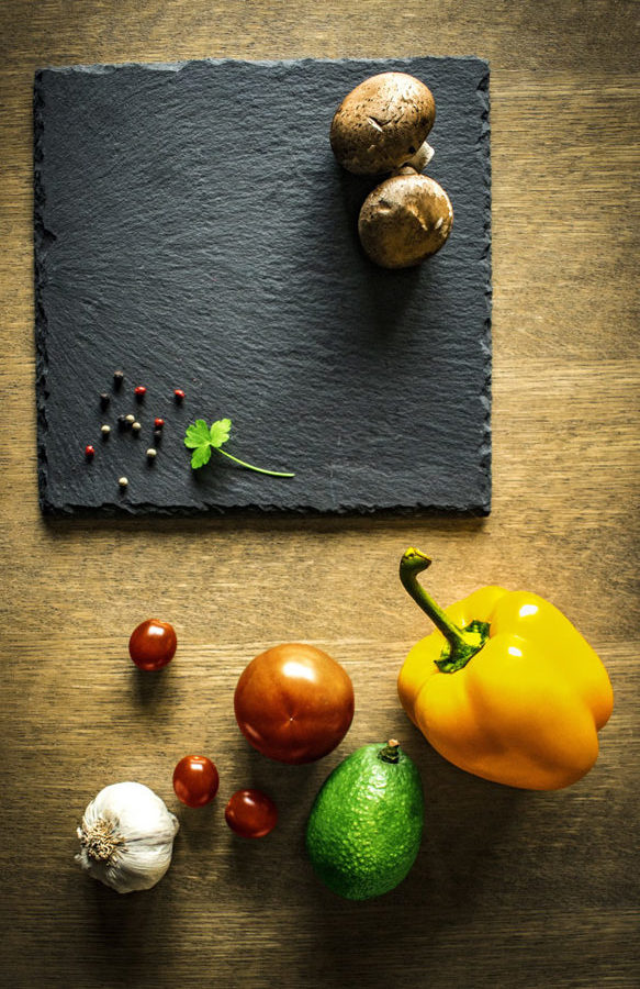 On a black slate board with mushrooms, peppercorns, and a parsley leaf sits above a table holding vibrant yellow bell pepper, avocado, tomato, cherry tomatoes, and a garlic bulb in Reno, Nevada.