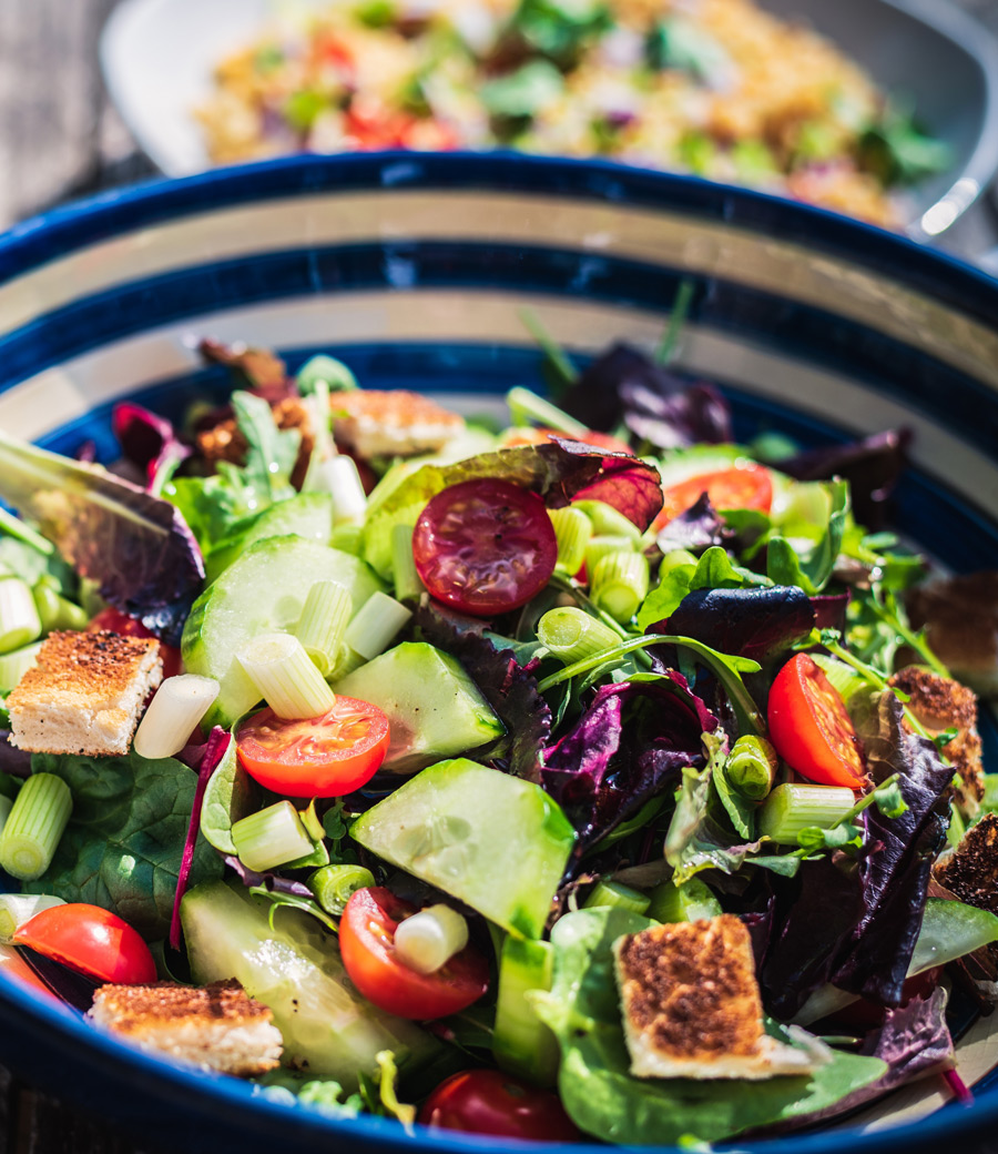 A colorful tossed salad in a blue and white bowl, featuring cherry tomatoes, cucumbers, croutons, and mixed greens, evokes the vibrant spirit of Reno. Blurred in the background is another bowl brimming with fresh salad—reminiscent of a peaceful moment at your favorite medical spa.