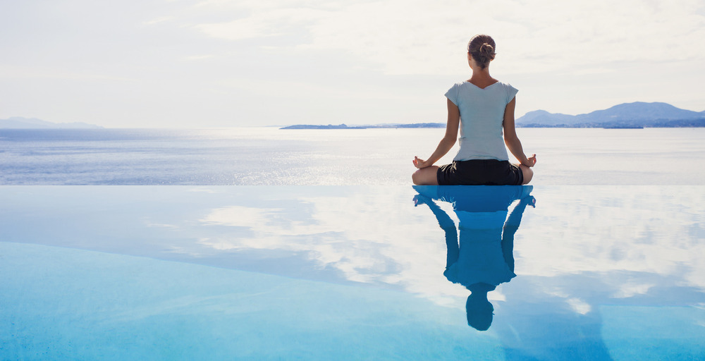 A person sits cross-legged on the edge of an infinity pool at a renowned medical spa, meditating while facing a calm ocean with a distant mountain range in the background, reminiscent of Nevada's serene landscapes.