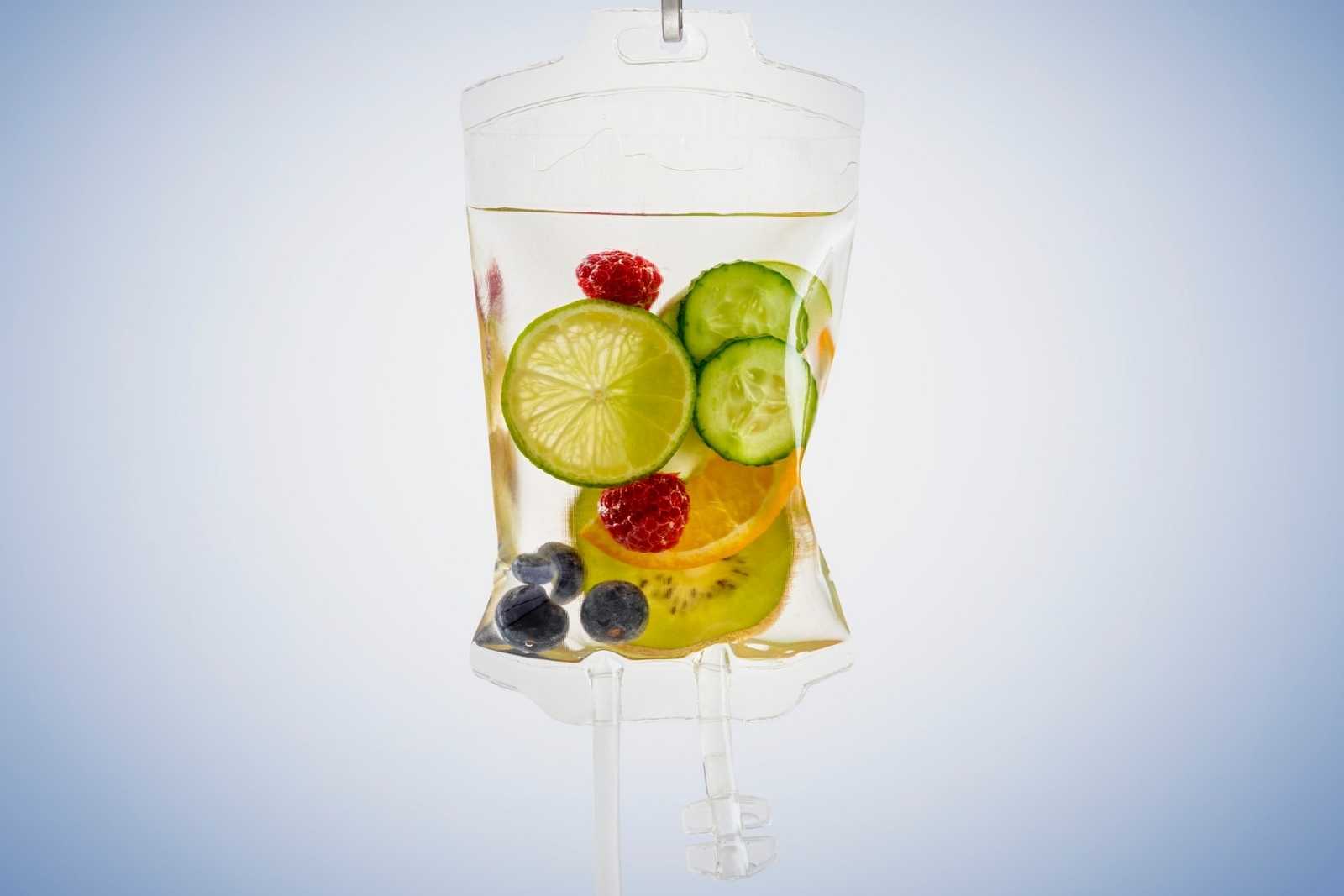 A transparent IV bag filled with slices of lime, cucumber, kiwi, and berries is set against a light blue background, evoking the refreshing treatments of a medical spa in Reno, Nevada.