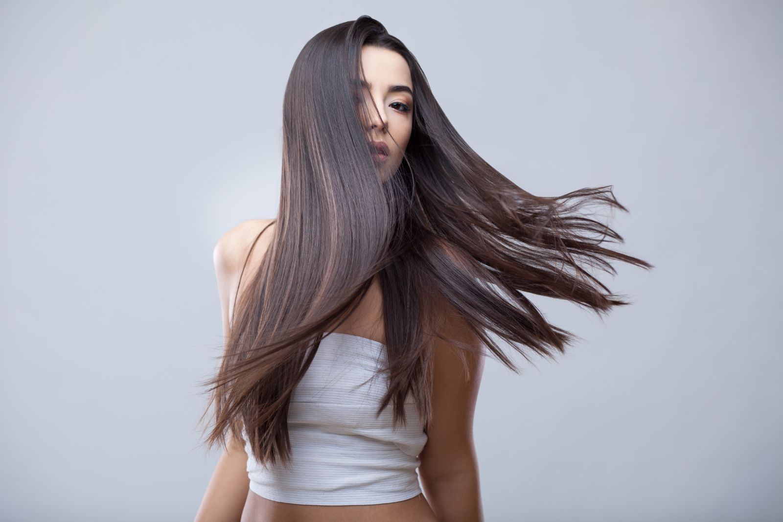 Person with long, straight brown hair wearing a white strapless top, her hair swinging to the side against a gray background, radiates an effortless elegance akin to the refined atmosphere of an aesthetics med spa in Reno.