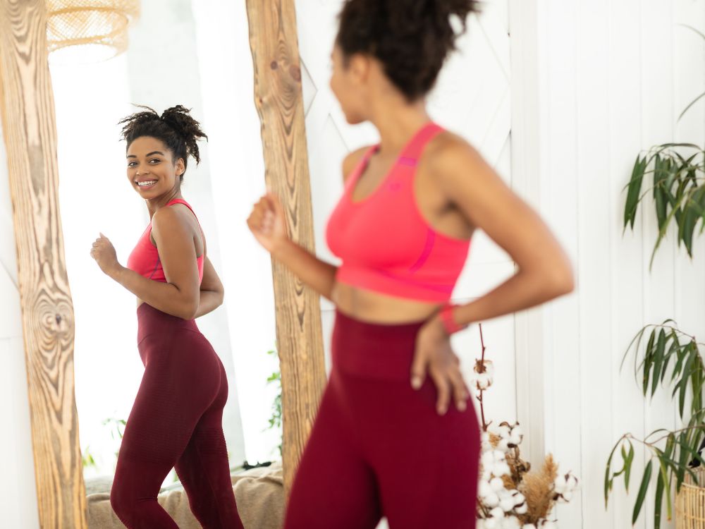A woman in activewear smiling at her reflection in a mirror while jogging in place.