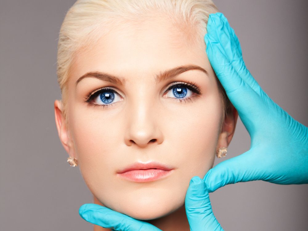 Two hands in blue gloves framing a woman's face with a neutral expression and direct gaze.