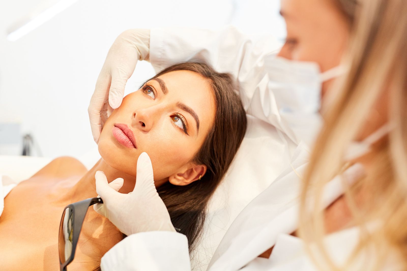 A dermatologist in gloves examines the face of a young female patient in a clinical setting.