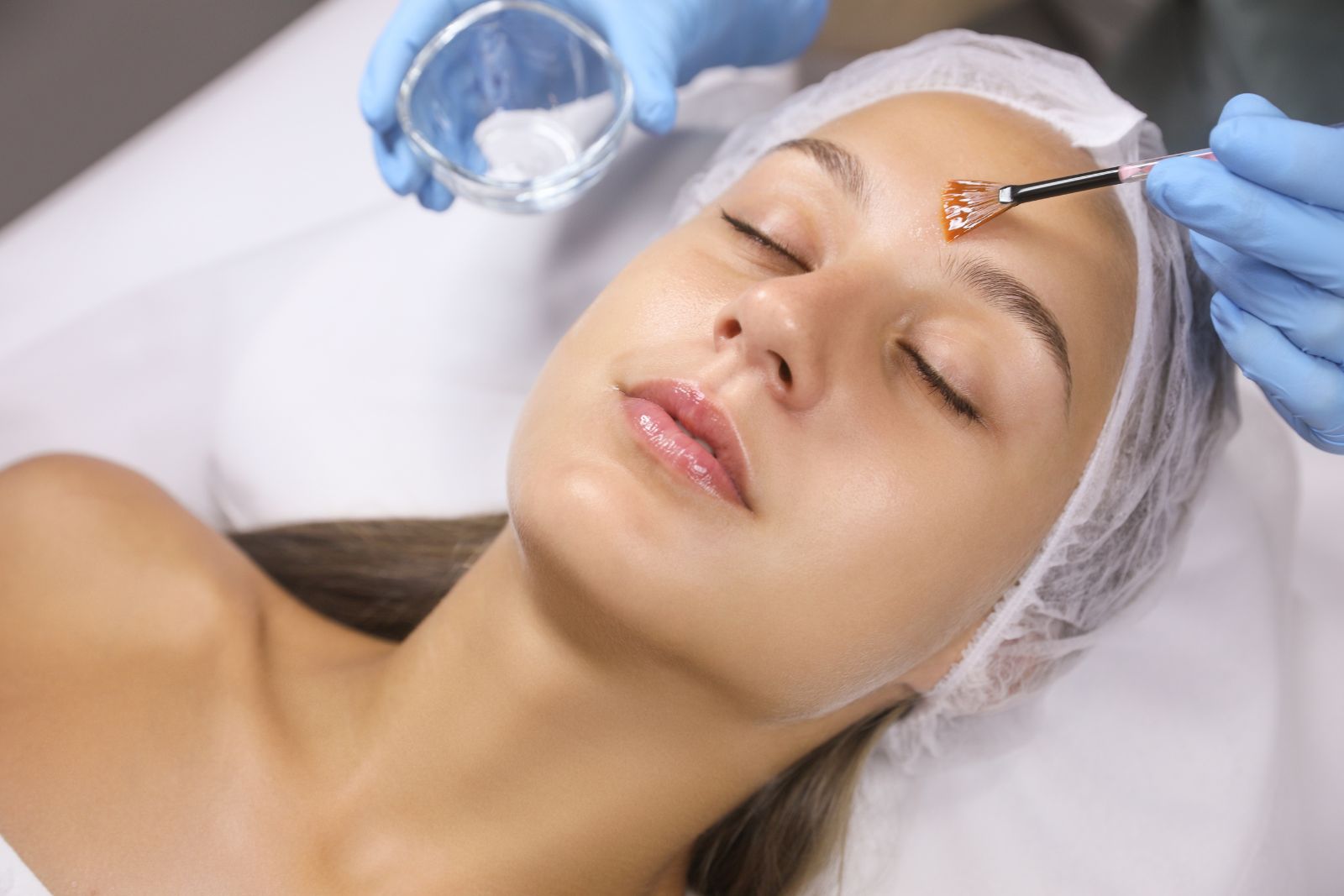 A woman receiving a facial treatment with a brush, lying down with her eyes closed, as a professional in gloves applies a product to her face.