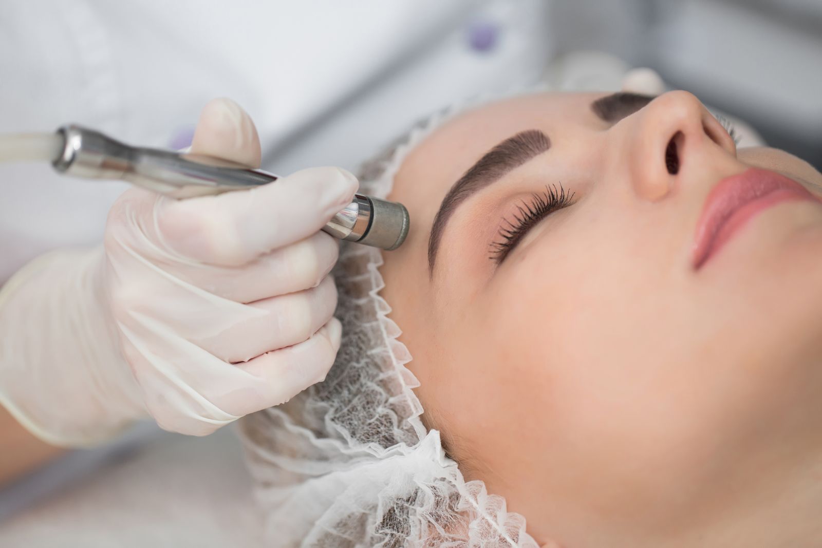 Aesthetician performing a facial treatment on a woman using a specialized machine in a clinical setting.