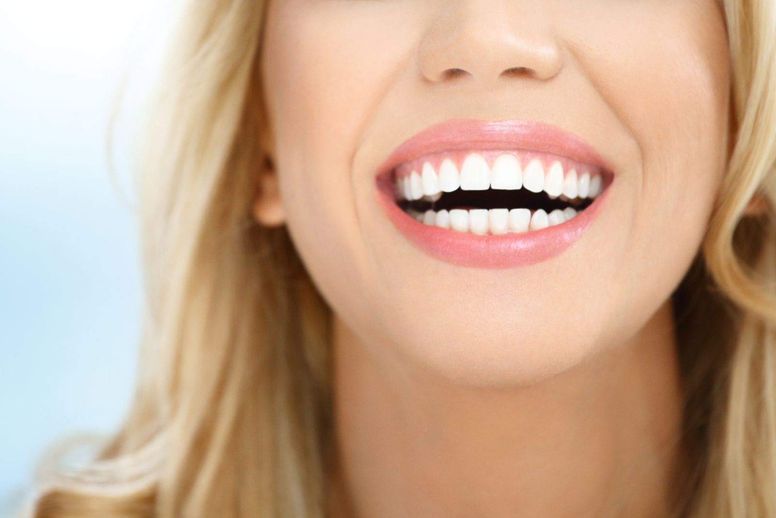 Close-up of a smiling woman with blonde hair, showing white teeth, partial view of her face against a light blue background.