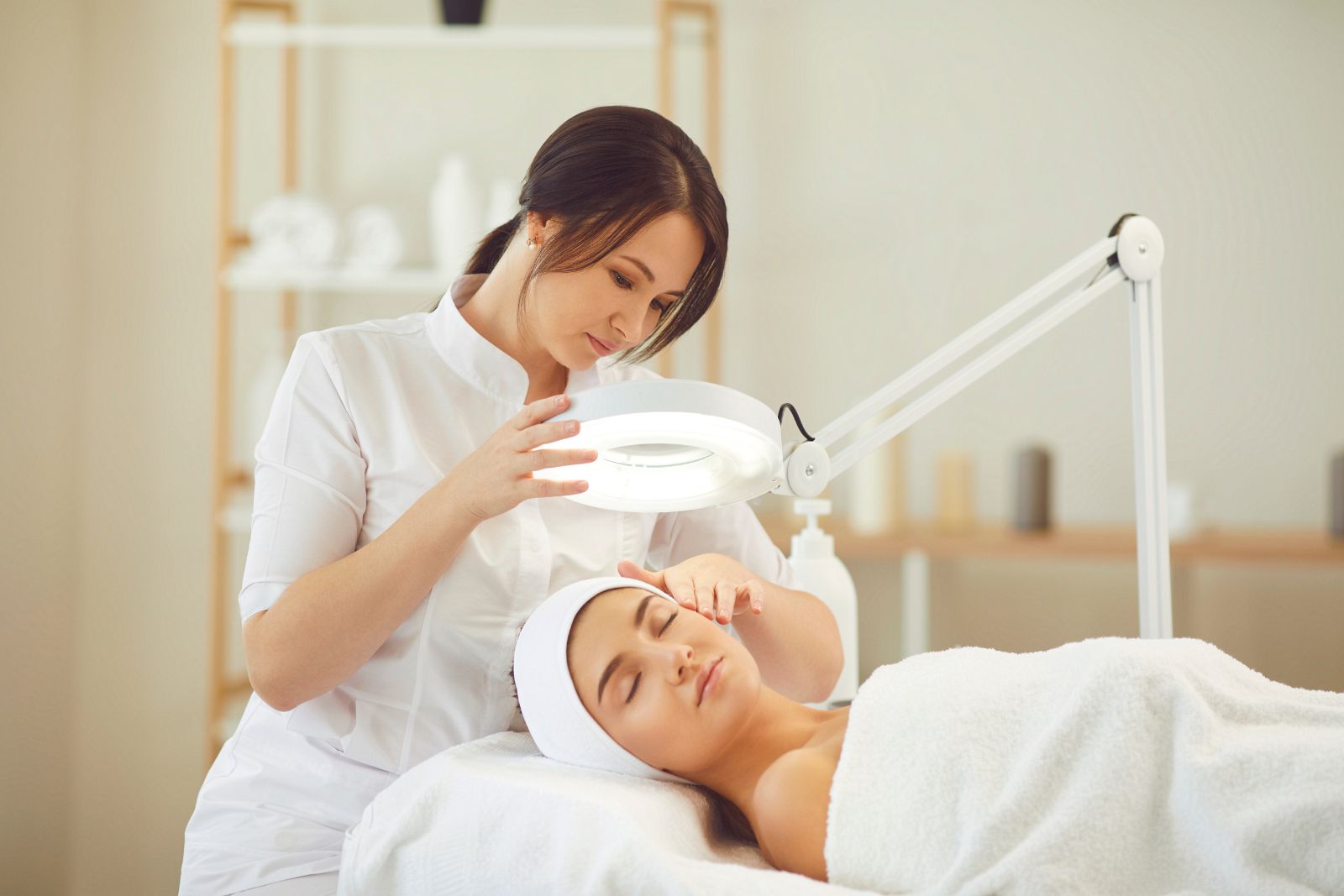 A female beautician using a magnifying lamp to examine a female client's skin during a facial treatment in a serene spa setting.
