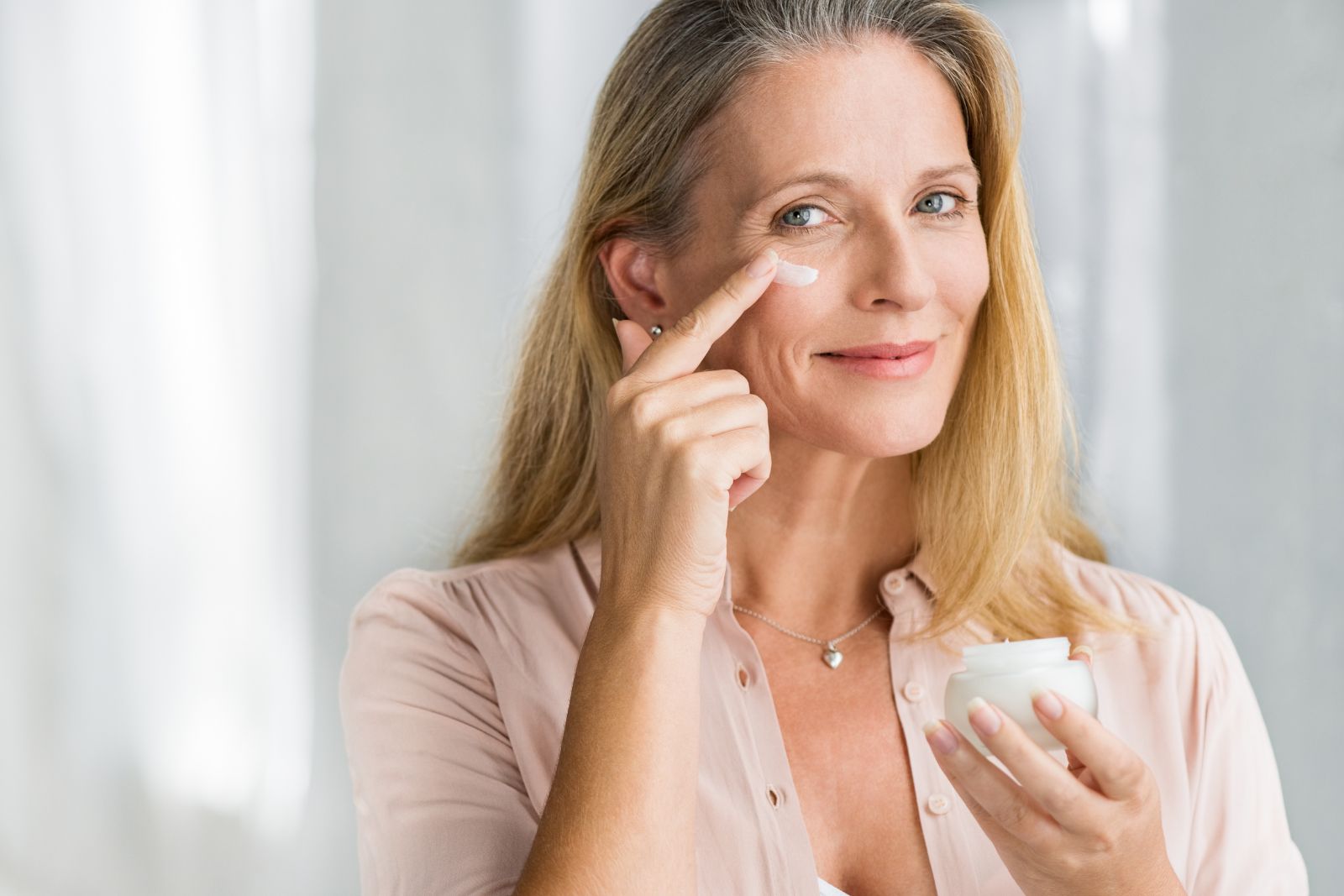 Mature woman applying facial cream under her eye, smiling gently, in a softly lit room.