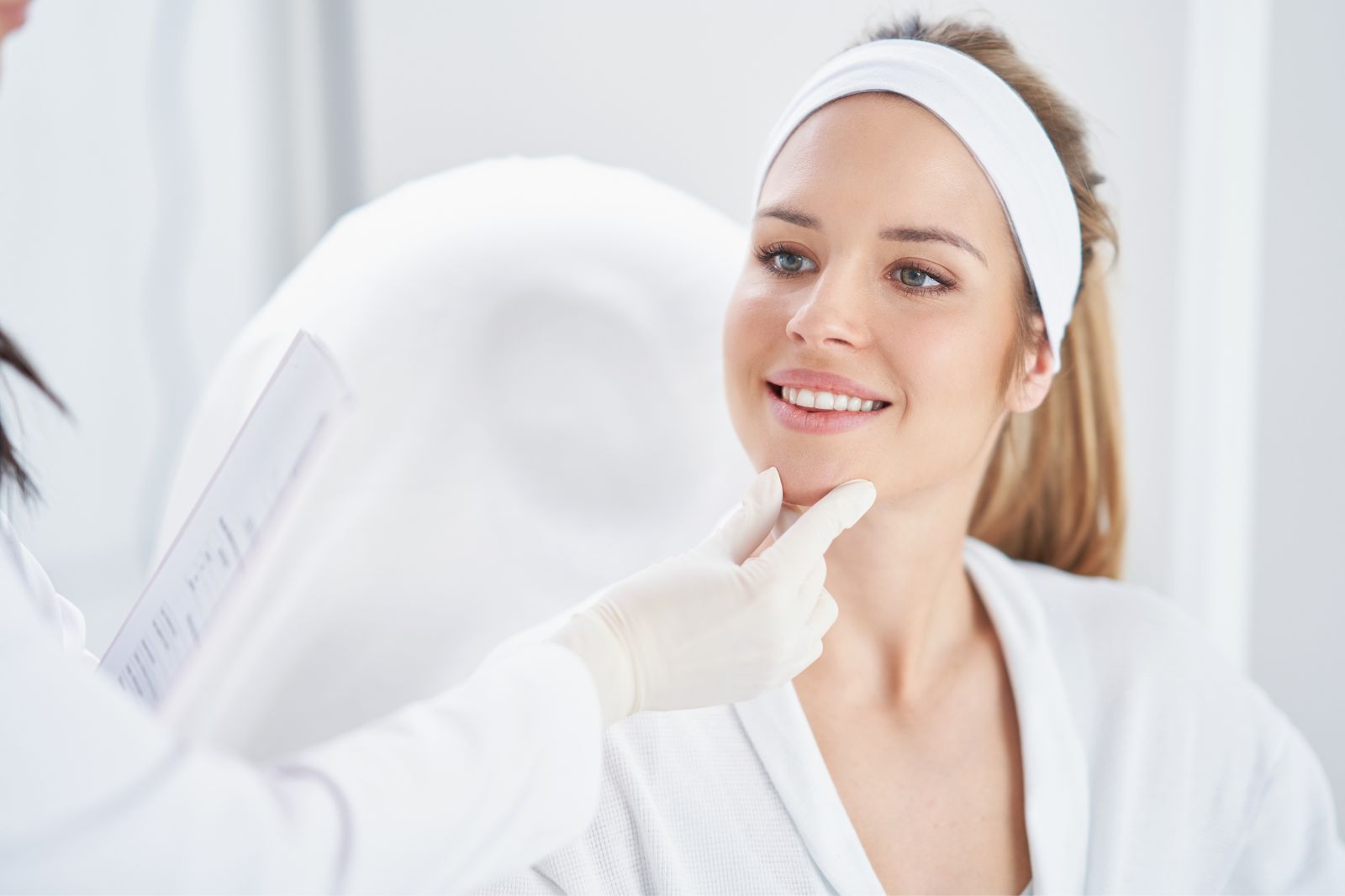 A smiling woman with a headband sits in a clinic, having her chin examined by a healthcare professional wearing gloves.