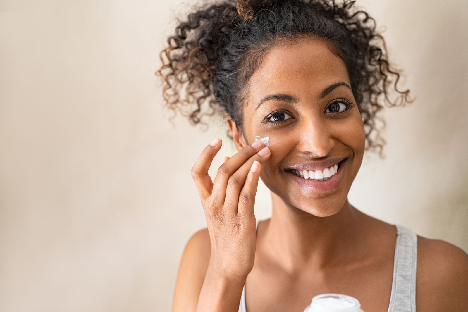 A person with curly hair applies cream to their face while smiling and holding a container in the other hand.