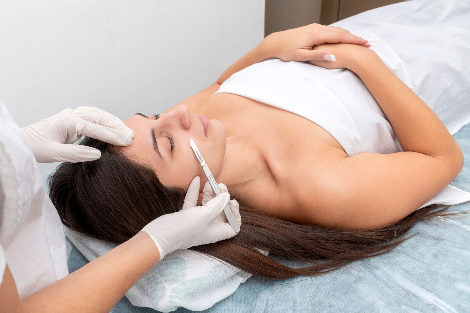 A person lying down with eyes closed while another person, wearing gloves, uses a surgical scalpel to perform a dermaplaning procedure on their face in Reno, NV.