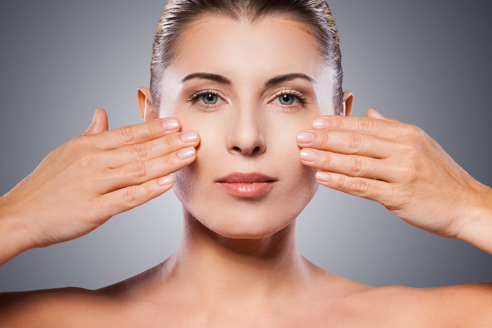 A person with fair skin and slicked-back hair holds their hands up to the sides of their face, showcasing a skincare routine. The background is a plain, gradient grey.