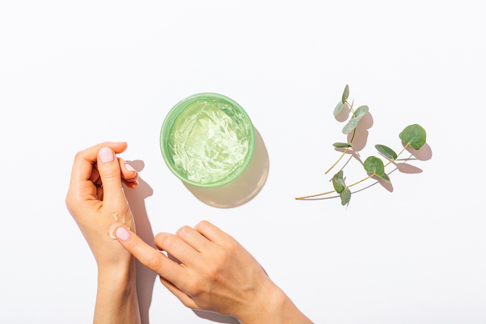 Hands applying a green gel from a jar, with eucalyptus leaves placed nearby on a white background.