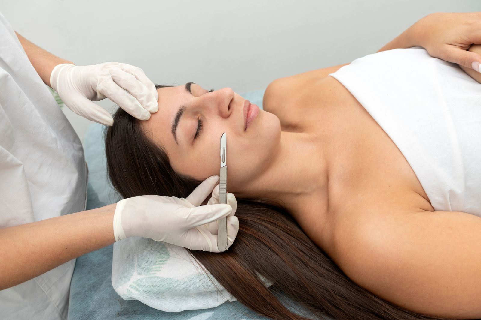 A person lying down with eyes closed receives a facial treatment with a scalpel from a gloved professional.