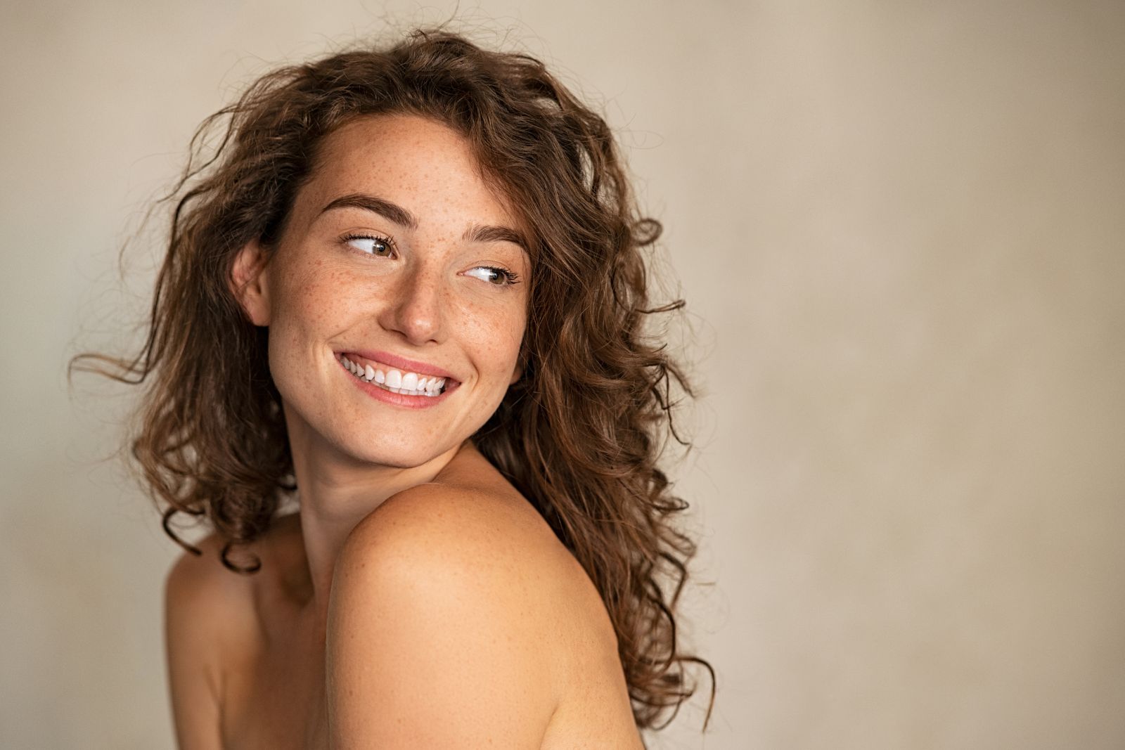 A smiling woman with curly brown hair and freckles looks to her left against a neutral background.