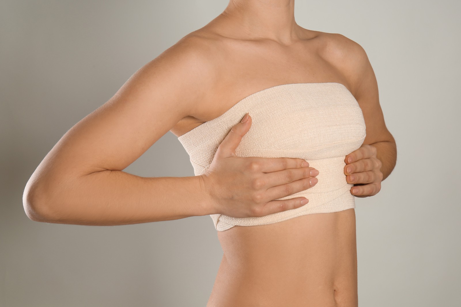 A person wrapping a beige bandage around their chest, covering the upper torso. The background is plain grey.