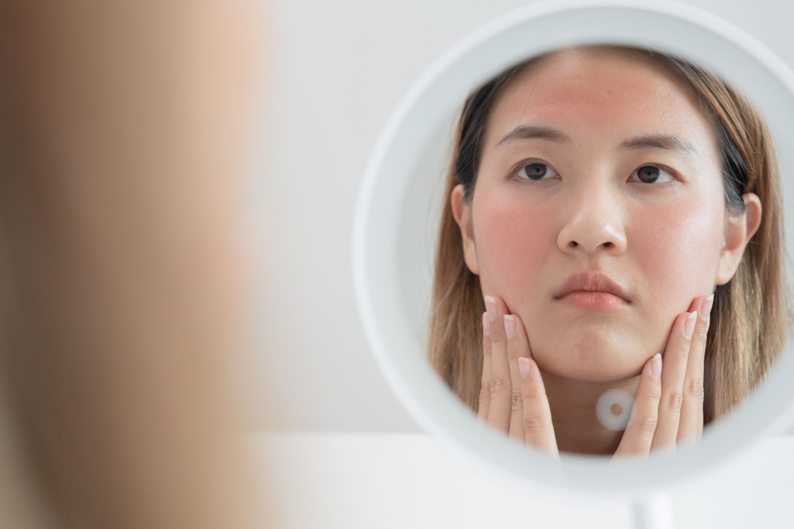 Person with a concerned expression looking at their reflection in a mirror, touching their red, irritated cheeks with both hands.