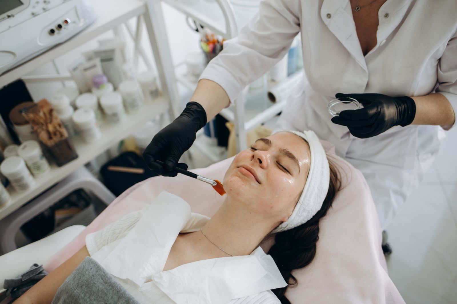 A woman receiving a facial treatment in a clinic, lying down with her eyes closed, as a professional applies a product to her face with a brush.