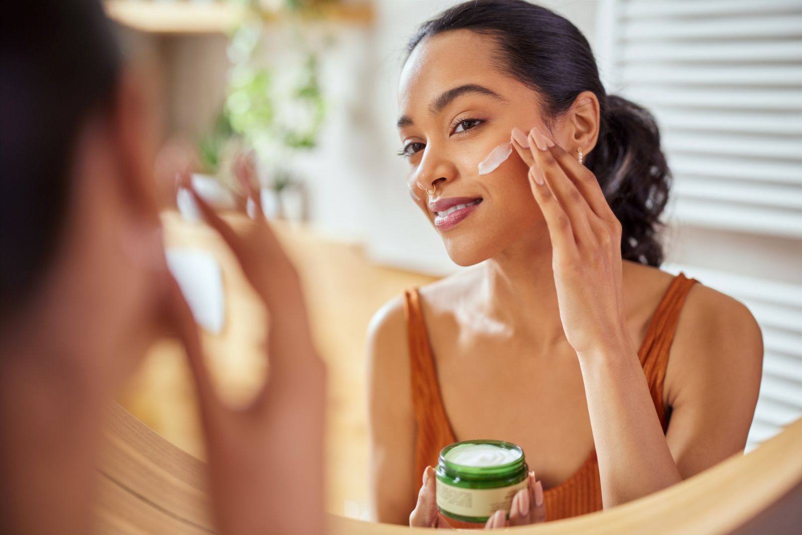 A woman applies skincare cream to her face while looking into a mirror, holding an open green jar of product in her left hand.