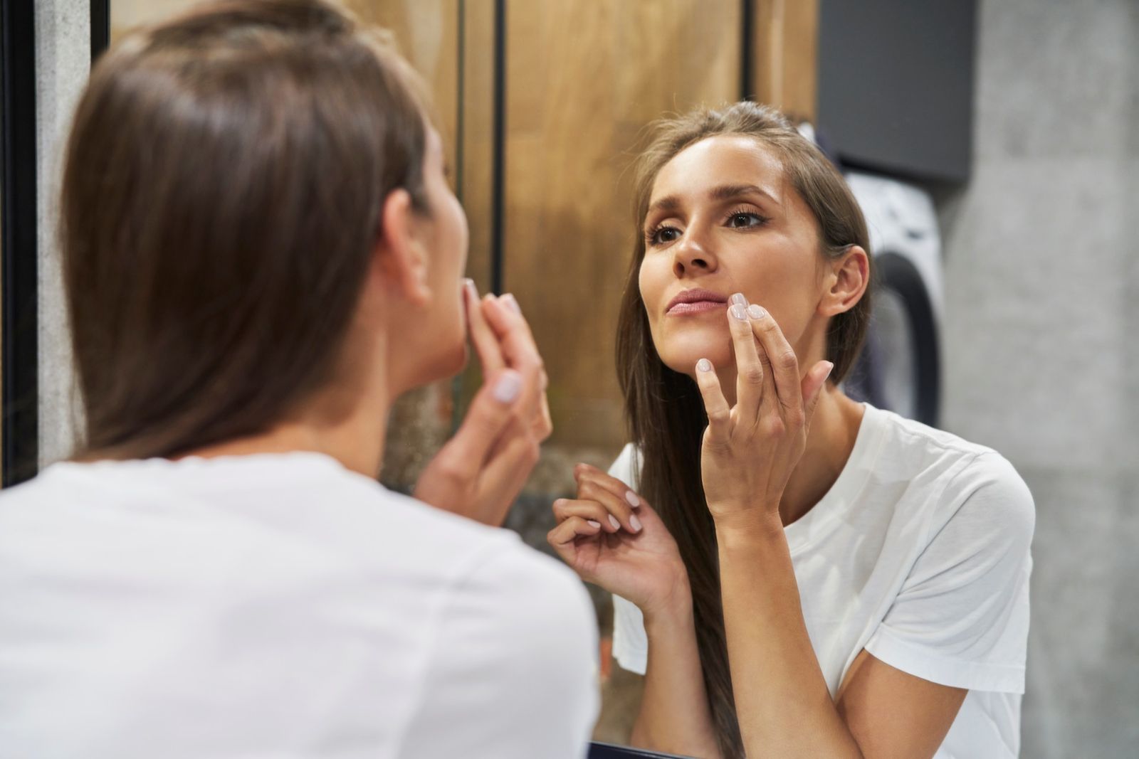 A woman in a white t-shirt looks at her reflection in a bathroom mirror while touching her face.