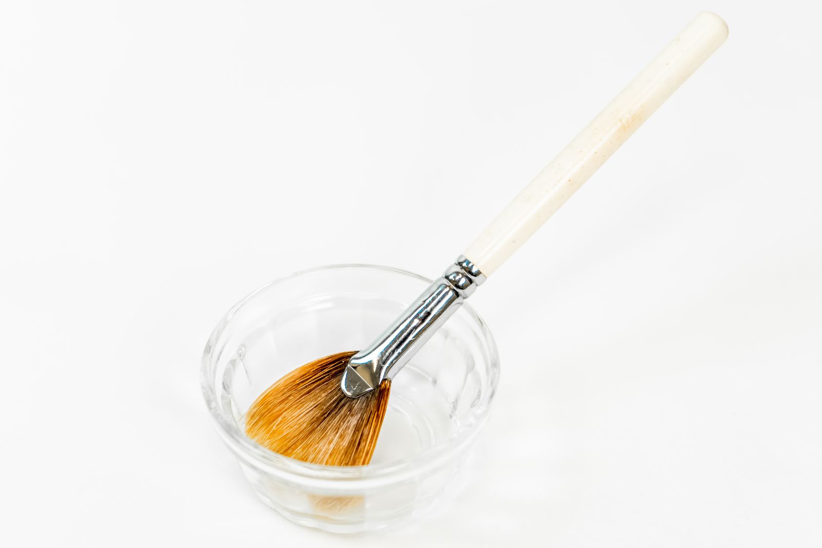 A paintbrush with a white handle rests inside a small, clear glass bowl containing a liquid, much like the tools used for applying a chemical peel on back treatments.