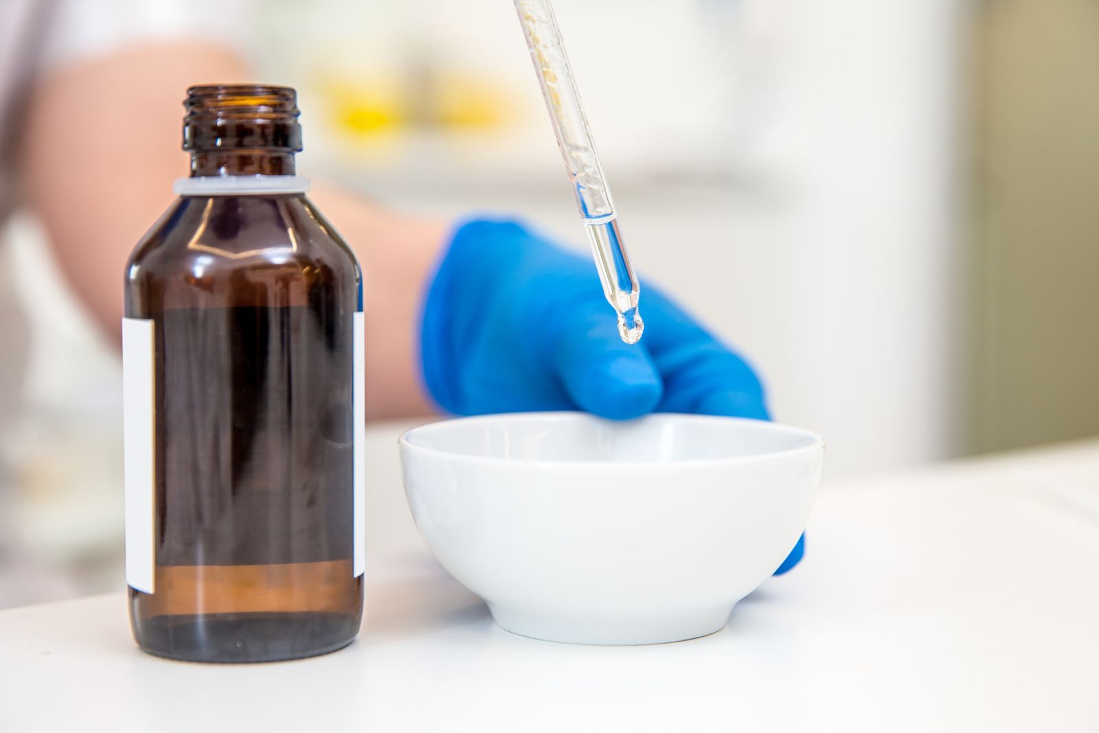 A person with a blue glove uses a dropper to add liquid into a white bowl next to a brown bottle on the counter, preparing for a chemical peel on back.