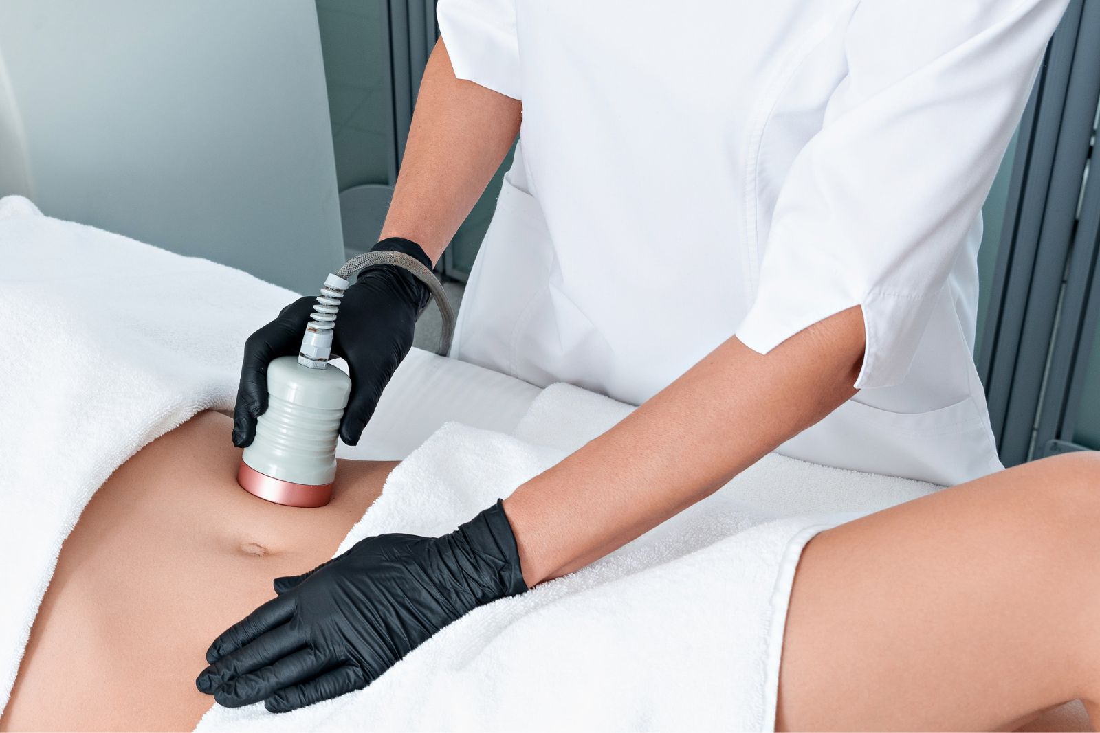 A person in black gloves performs an Ultrashape Fat Reduction procedure on a client's abdomen using a specialized device. The client is covered with a white towel.