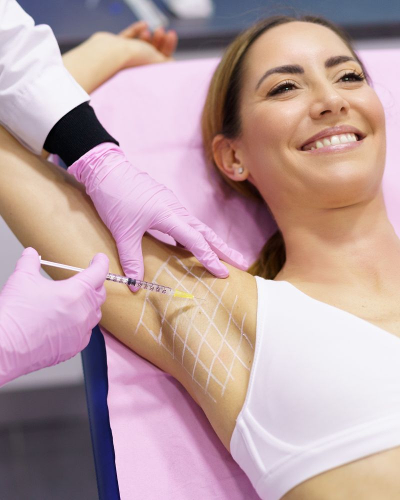 A woman lying down, smiling, while a medical professional wearing pink gloves administers armpit botox into her marked underarm area.