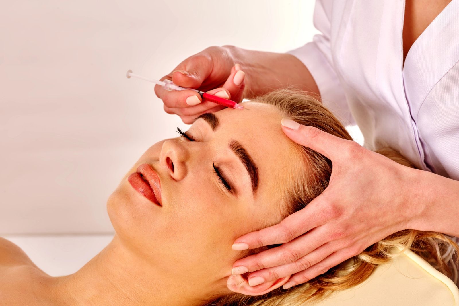 A person with closed eyes receives dermal filler injections in the forehead from a practitioner wearing a white coat.