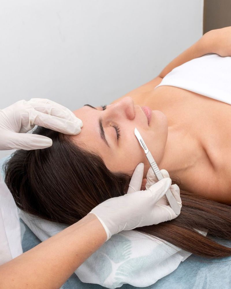 A person lies down while another, wearing gloves, uses a small scalpel to perform a dermaplaning facial treatment.