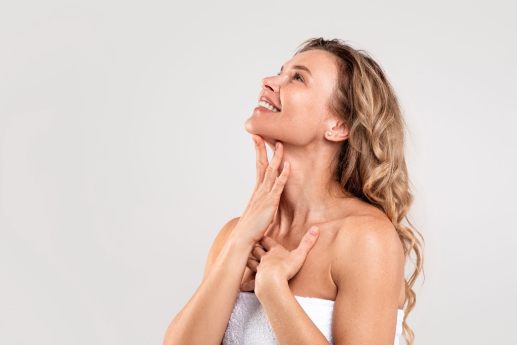 A woman with long, wavy blonde hair wrapped in a white towel looks upwards while touching her neck with both hands, perhaps contemplating thyroid injections, against a plain background.