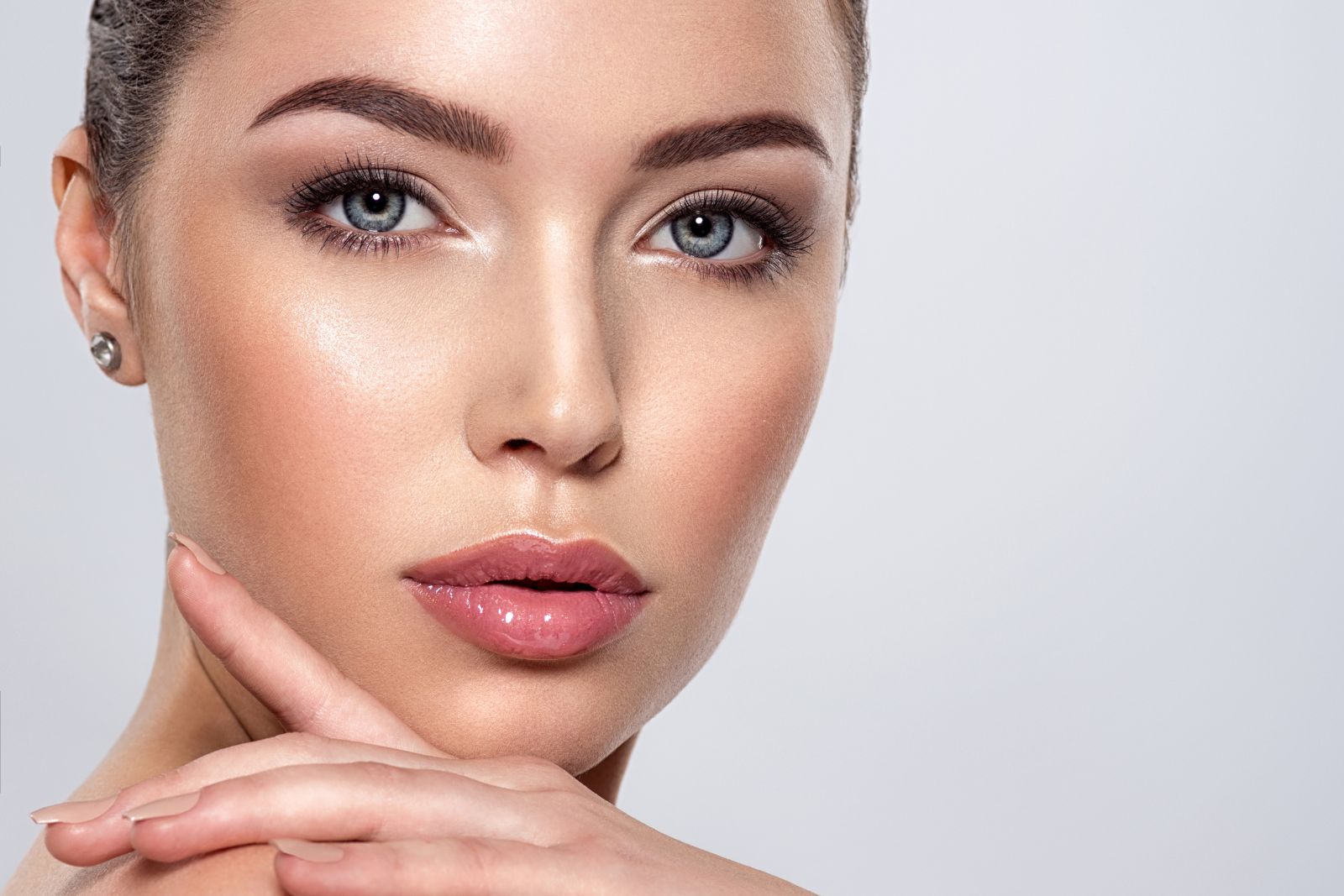 Close-up of a woman with light skin, smooth complexion from Glutathione Collagen and Vitamin C, light blue eyes, and pink lips. She's touching her chin with her index finger. She has neatly groomed eyebrows and wears stud earrings.