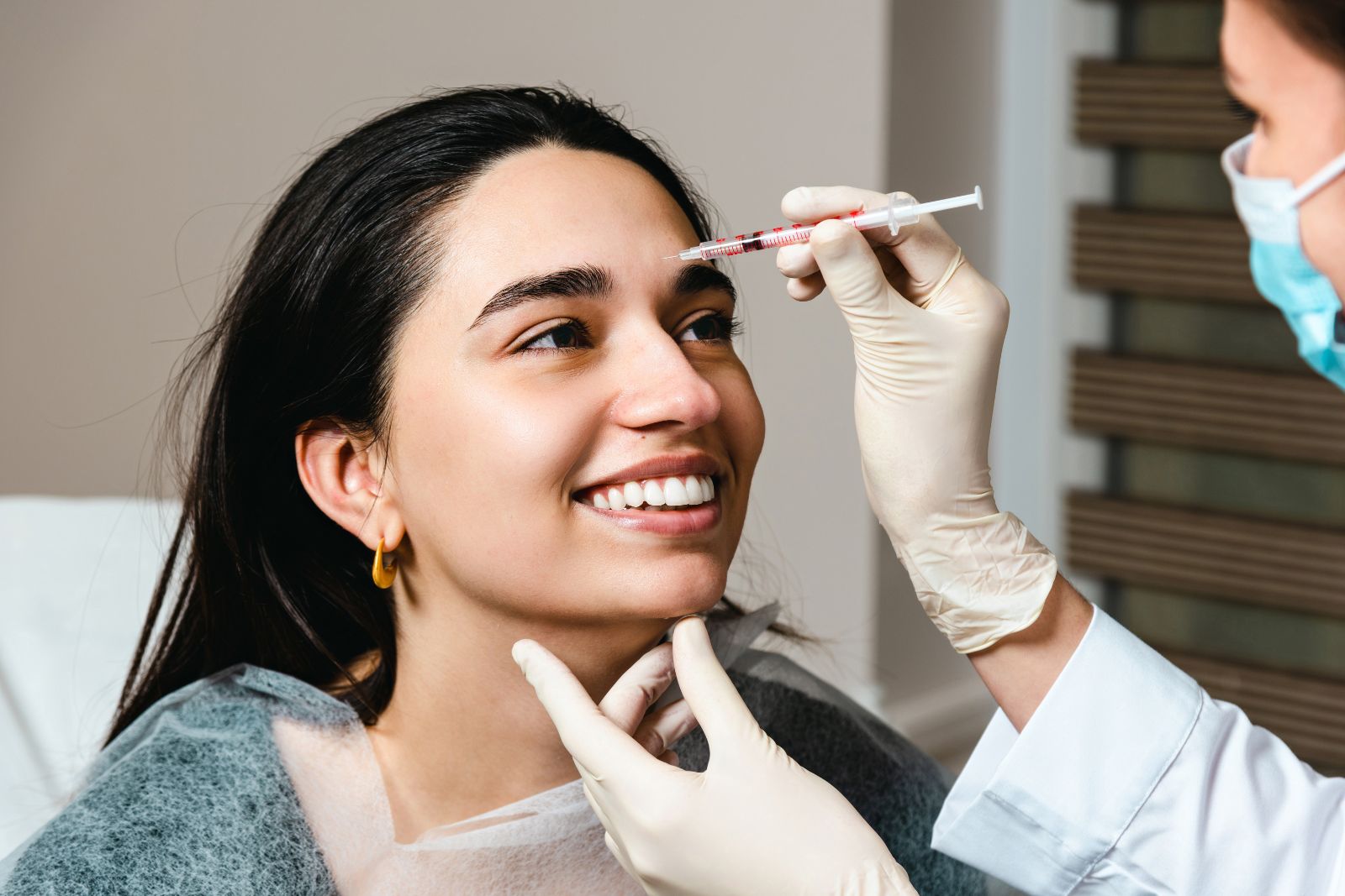 A woman smiles as a healthcare professional wearing gloves and a face mask administers a jeuveau syringe injection to her forehead.