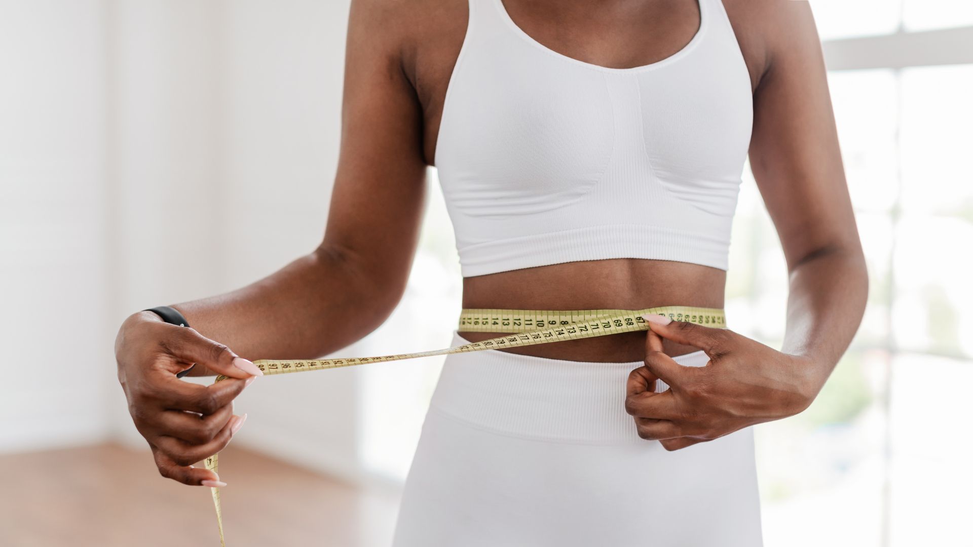 In a bright room, a person wearing a white sports bra and leggings measures their waist with a yellow measuring tape, perhaps preparing for their next Skinny Shot.