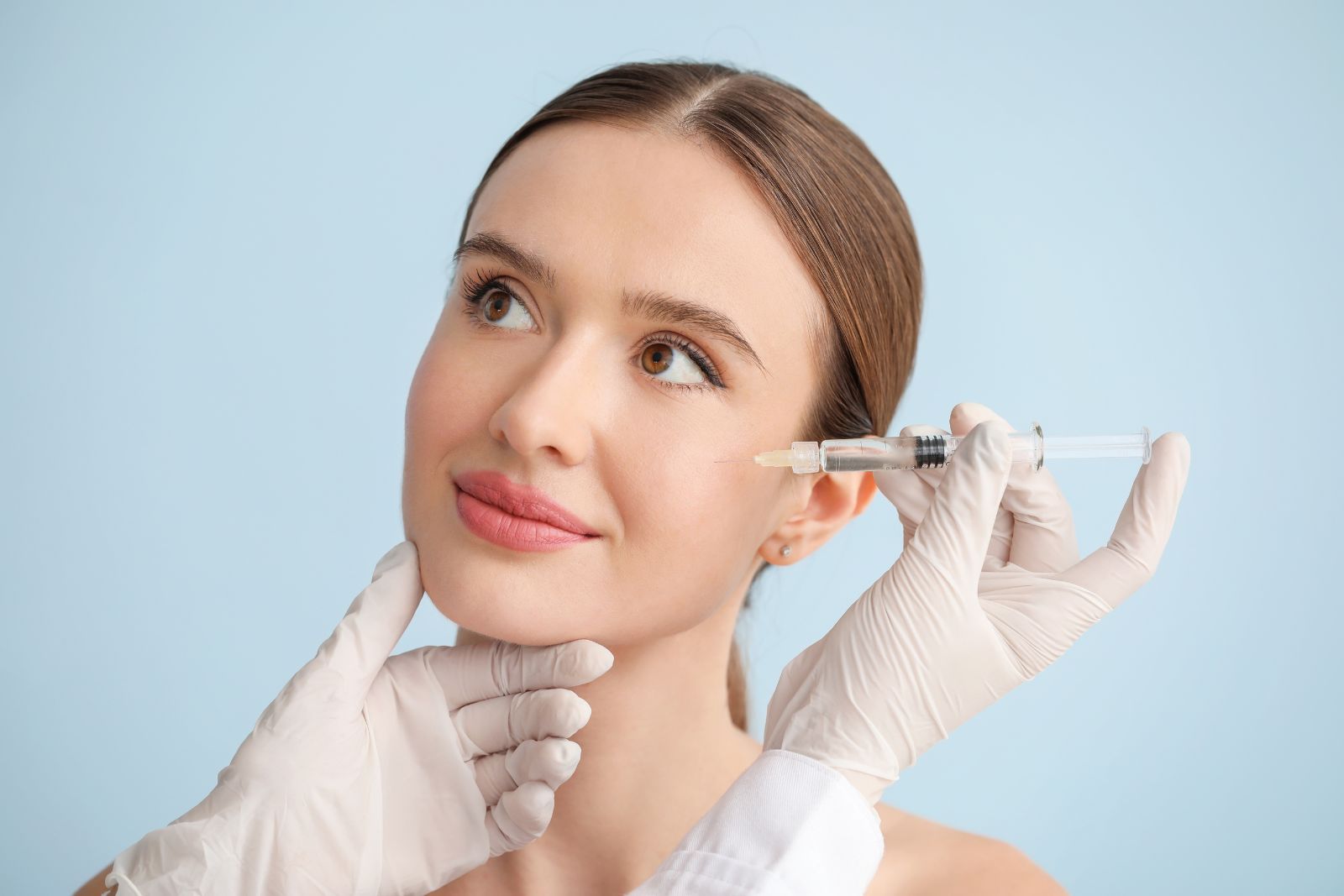 A person receiving a Jeuveau injection in the cheek from a healthcare professional wearing white gloves. Both are against a light blue background.