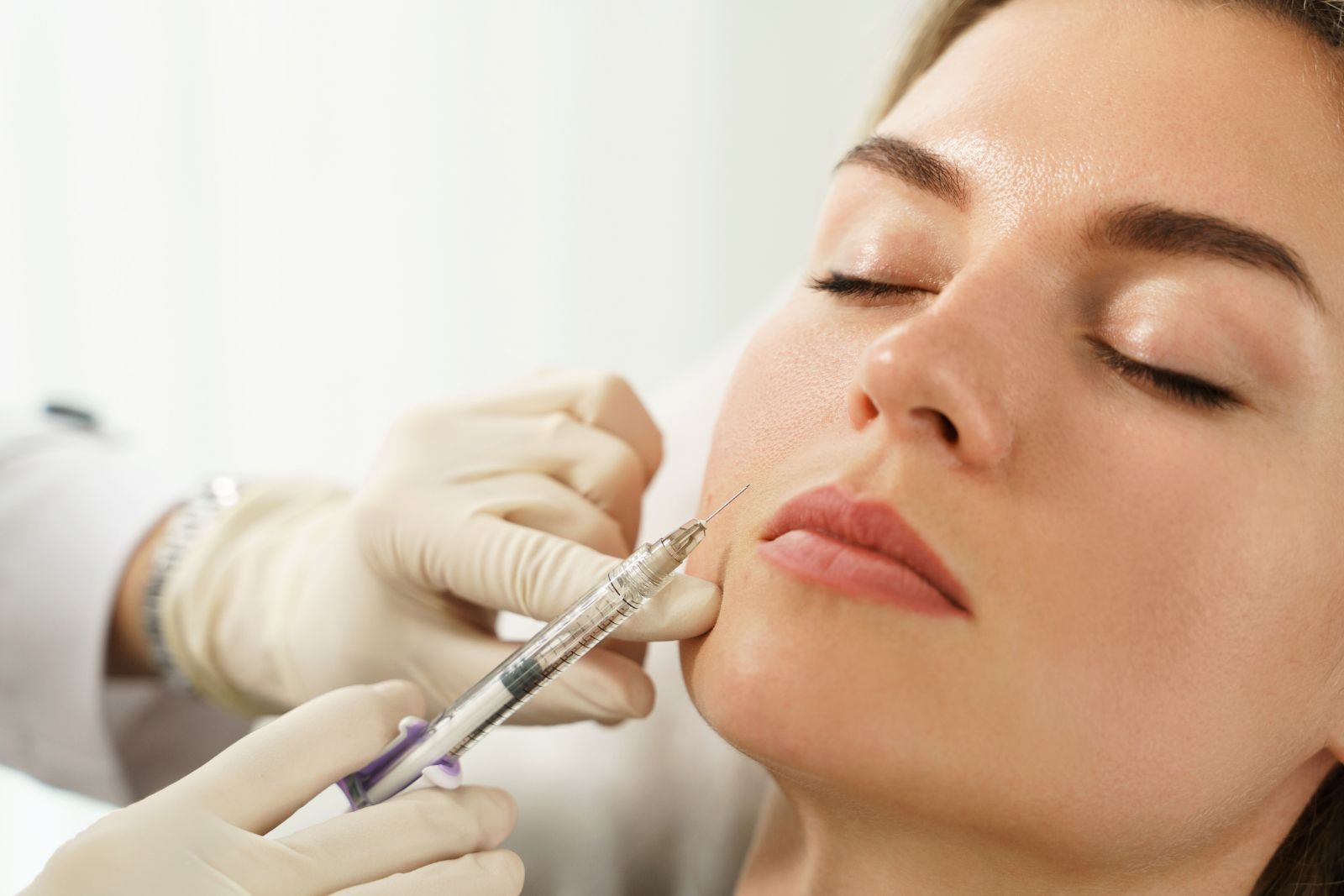 A close-up of a person's face with closed eyes receiving a Juvederm filler injection in the cheek from a medical professional wearing gloves.