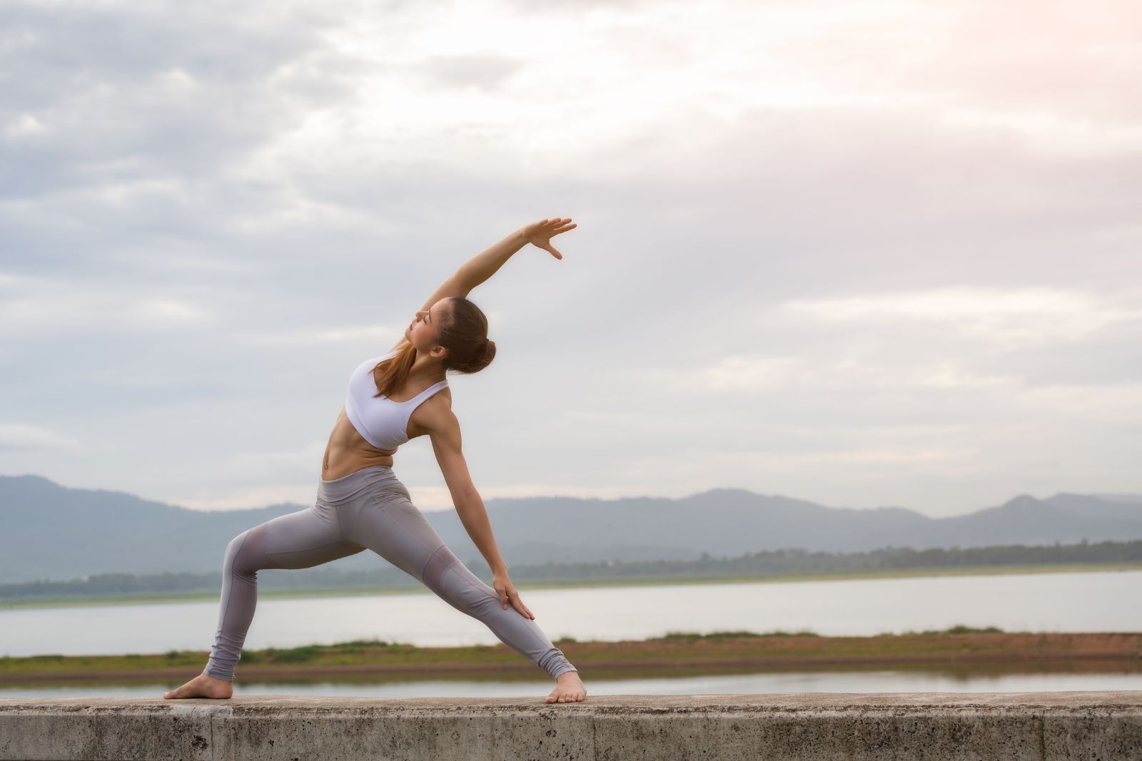 A person practicing yoga in a side angle pose on a concrete surface near a tranquil body of water, mountains framing the horizon—a perfect retreat for those seeking balance and serenity. Visit our aesthetics med spa to enhance your revitalization journey amidst nature's splendor.