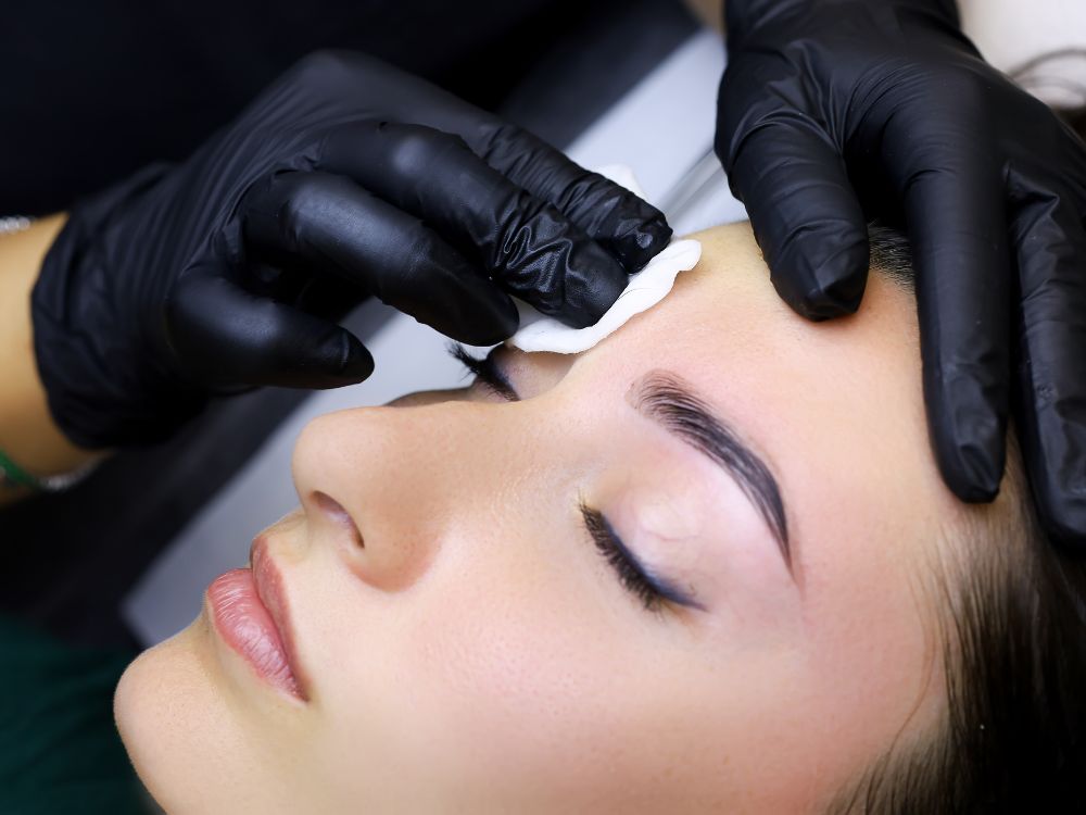 At our aesthetics med spa, a client enjoys a soothing brow treatment with eyes closed. Expert hands in black gloves gently apply a cotton pad to the eyebrow area, ensuring a serene and rejuvenating experience.