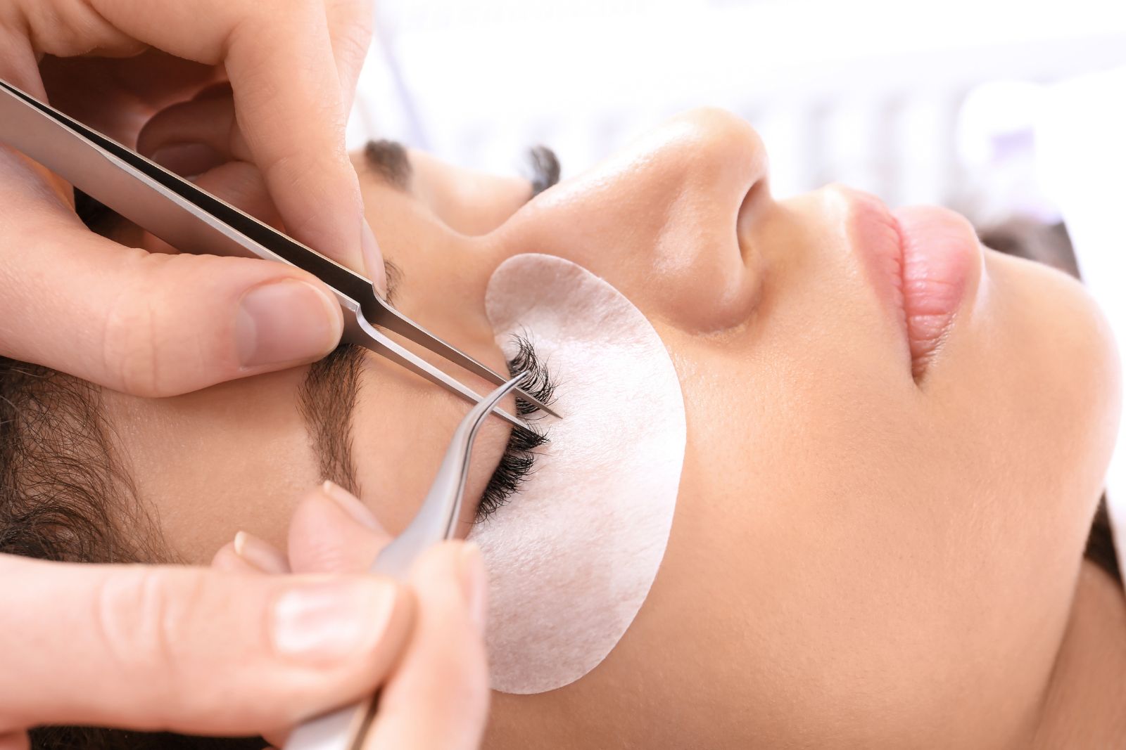 Close-up of a person at an aesthetics med spa receiving eyelash extensions, with tweezers applying lashes and a protective pad under the eye.