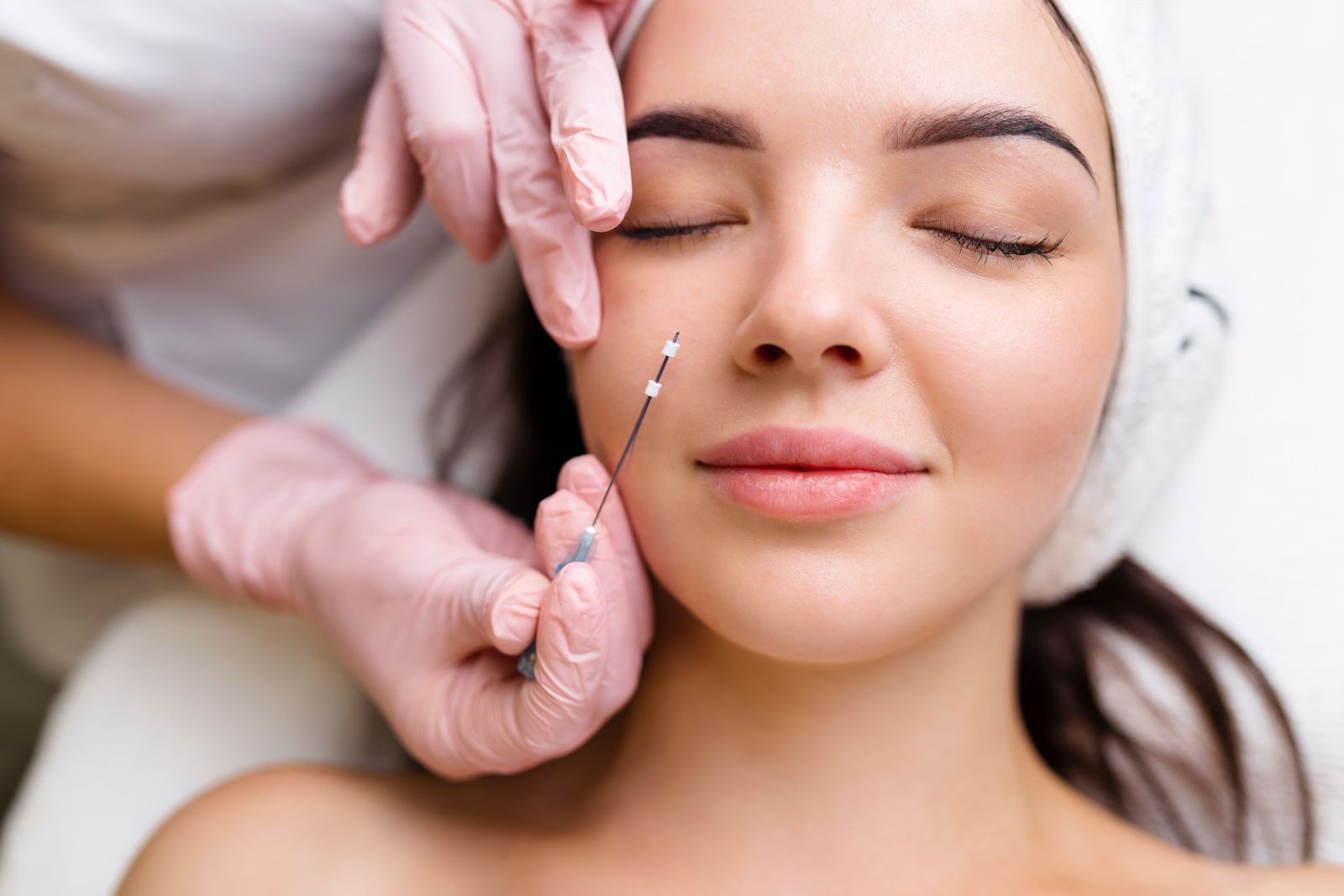 A person with closed eyes receives a facial injection from a gloved technician. The person is lying down and wearing a white hairband, opting for PDO Threads for under eyes to achieve a rejuvenated look.