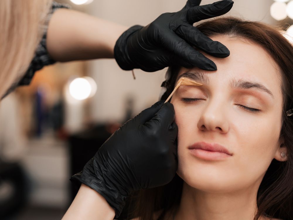 At a renowned aesthetics med spa in Reno, a person receives expert eyebrow shaping from a stylist wearing black gloves and skillfully wielding a wooden tool.