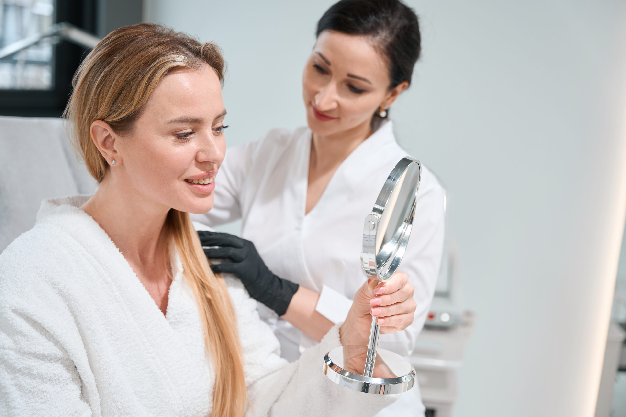 A woman in a white robe holds a handheld mirror, smiling as she admires the results of her PDO Threads for Neck procedure, while a professional in a white coat stands beside her in the clinical setting.