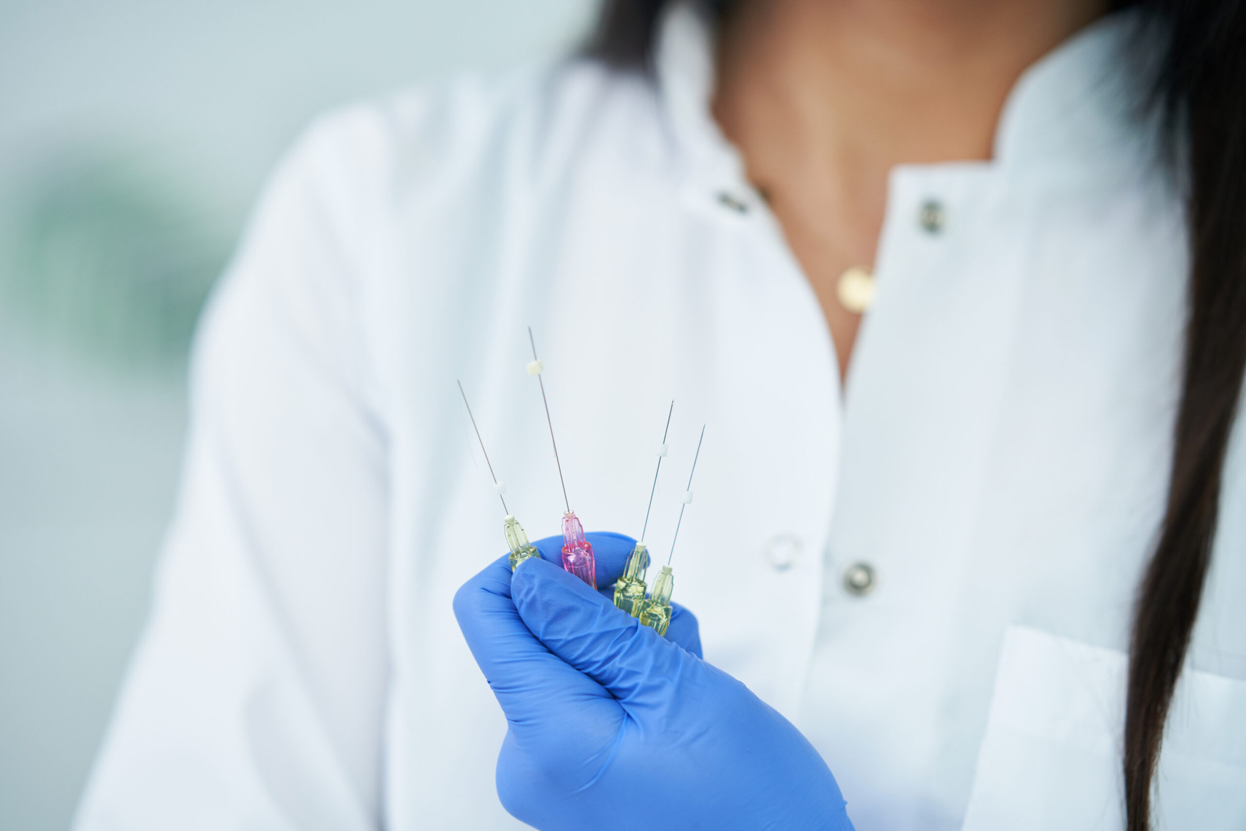 A person in a white lab coat and blue gloves carefully holds three syringes filled with colorful liquid, ready for a procedure involving PDO threads for jowls.