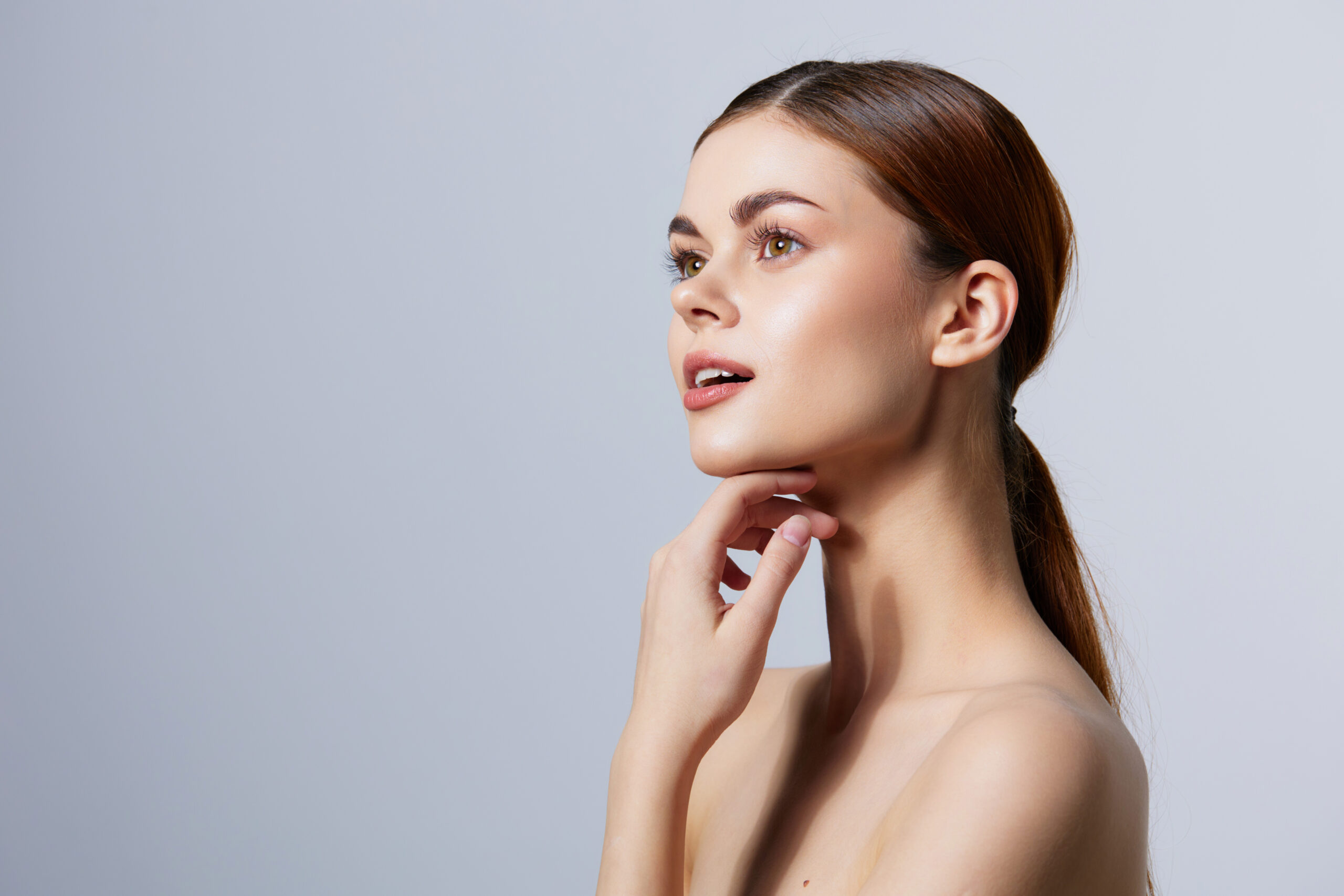 A woman with brown hair pulled back, looking upwards with a thoughtful expression, touches her chin against a plain background, perhaps contemplating the effects of PDO threads for jowls.