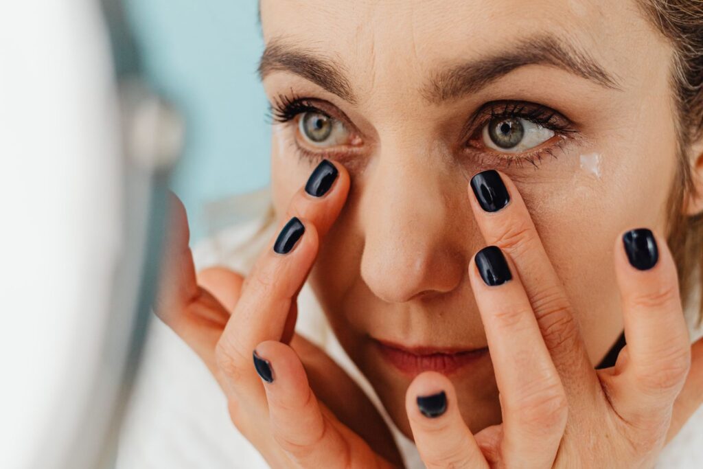 A person with dark nail polish applies cream under their eyes while looking in a mirror, considering the rejuvenating benefits of PDO threads for under-eye wrinkles.