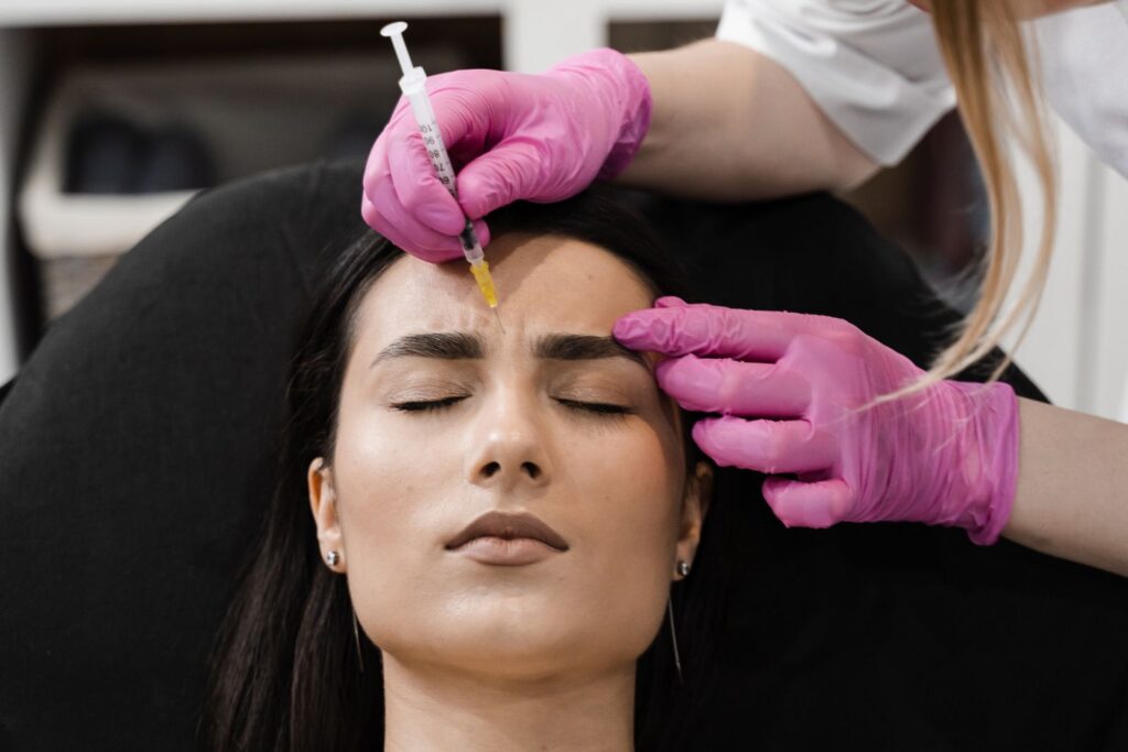 A person receives a forehead injection while lying down, administered by someone in pink gloves. Curious about types of dermal fillers? This treatment could be your answer.