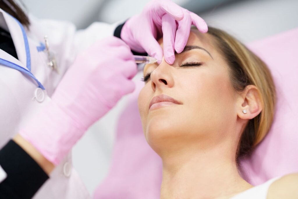 A woman lies down with her eyes closed as a person in pink gloves administers an injection to her face.