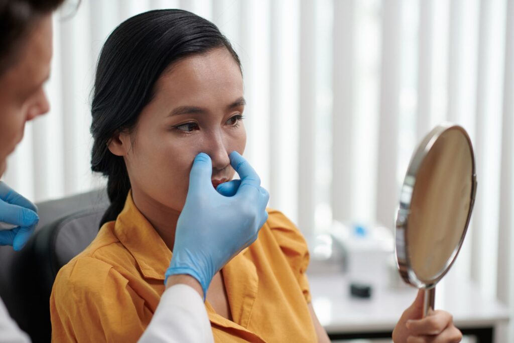 A woman in an orange shirt sits holding a mirror as a gloved doctor examines her nose in a medical office.