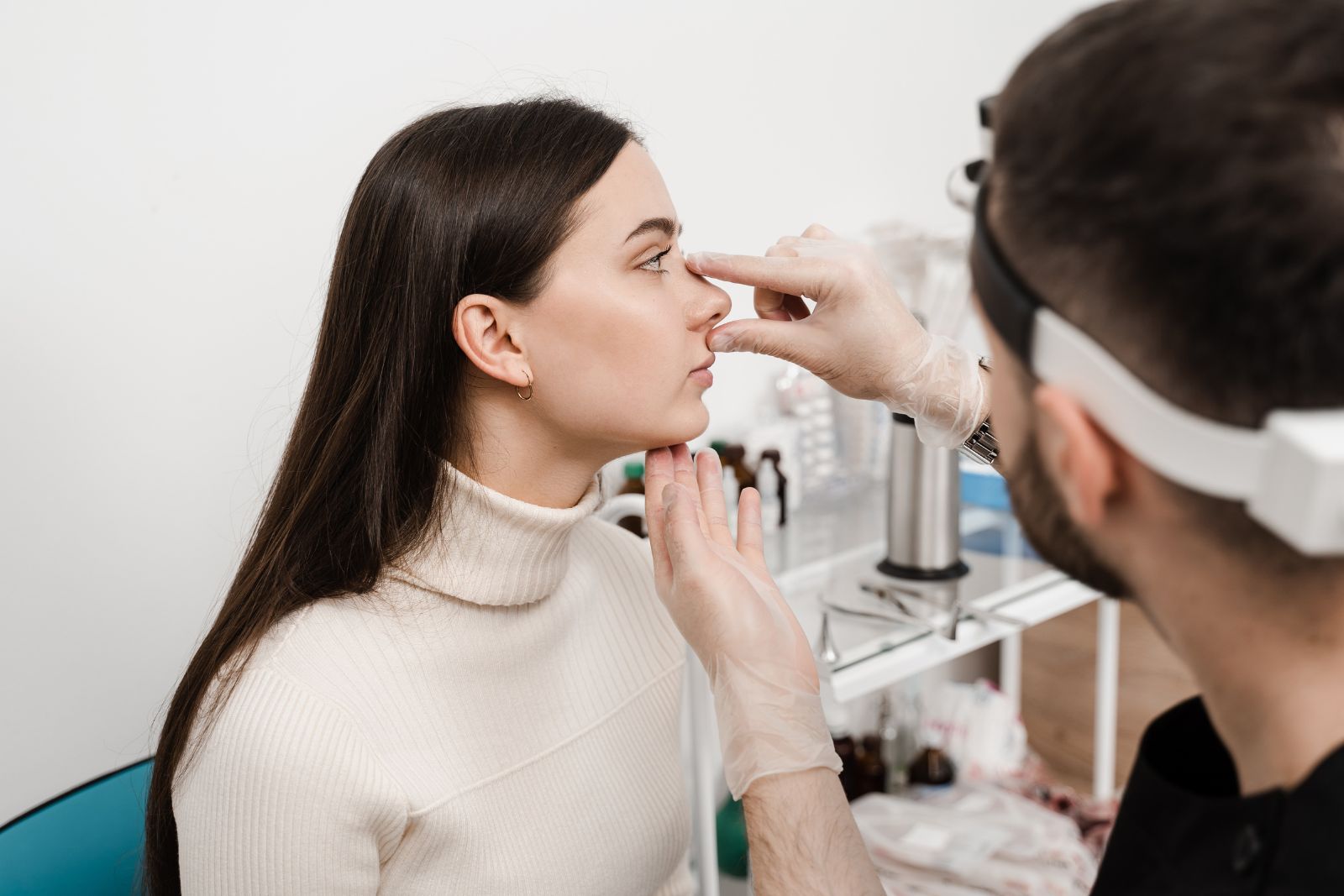 A doctor wearing gloves examines a woman's nose in a medical office. The woman is seated and looking straight ahead.